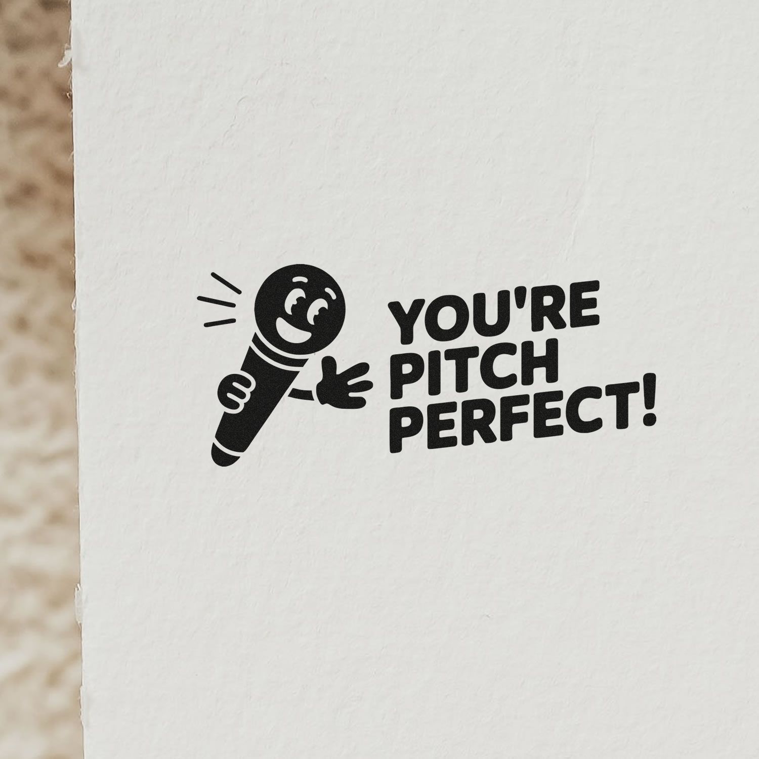 Black stamp and seal design with smiling microphone and Youre pitch perfect! on white paper.