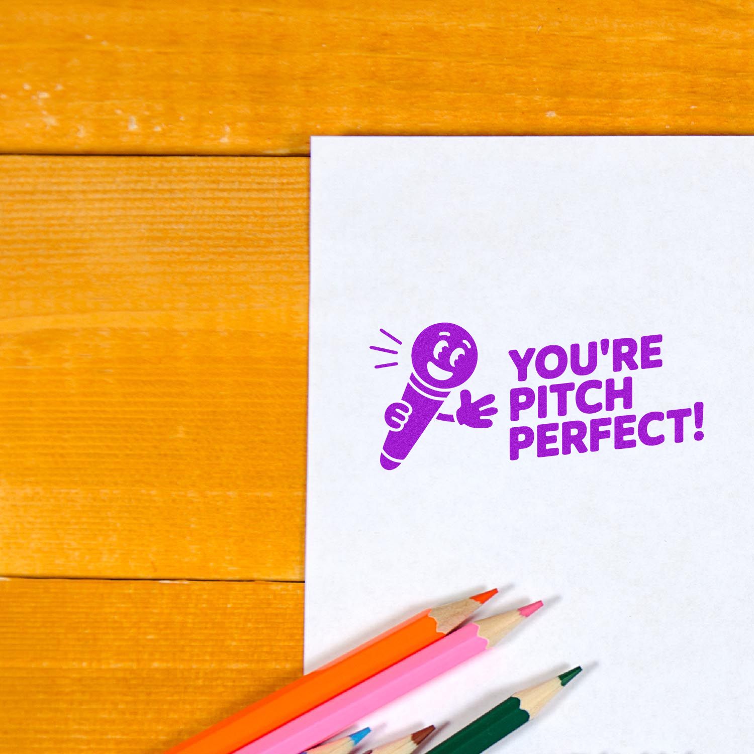 Purple YOURE PITCH PERFECT! stamp seal on white paper with orange, pink, and green pencils on a warm wood table.
