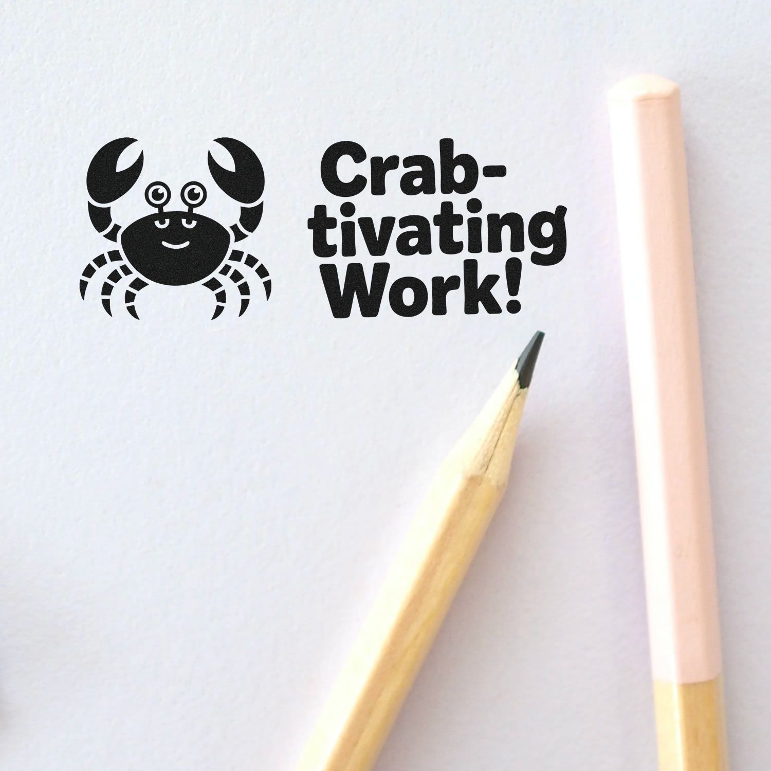 Black crab rubber stamp seal reading Crab-tivating Work! beside two pencils on white paper.