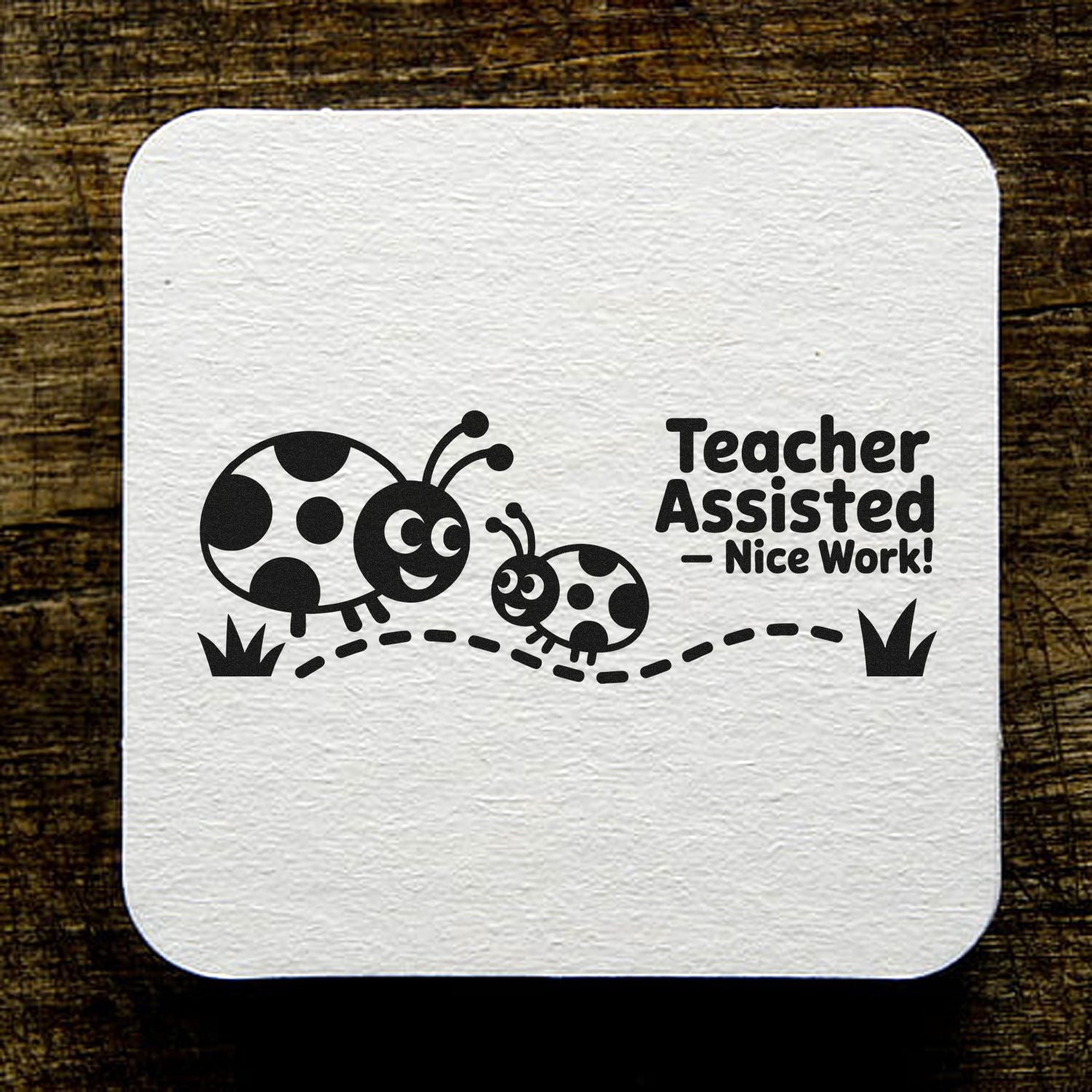 White rubber stamp seal with black ladybugs and Teacher Assisted - Nice Work! text on a wood background.