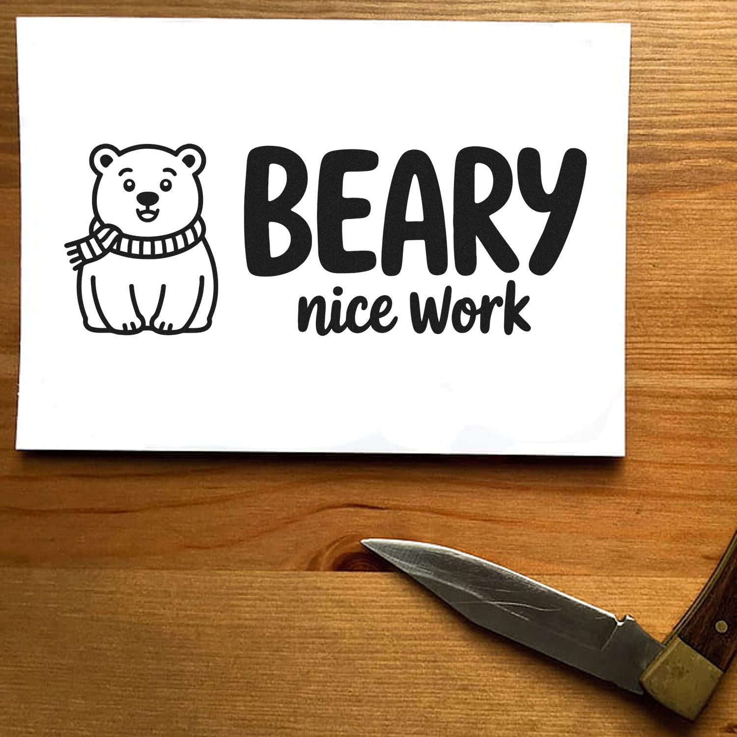 Black bear rubber stamp with scarf and BEARY nice work text on white paper beside a wood-handled knife on a wooden table.