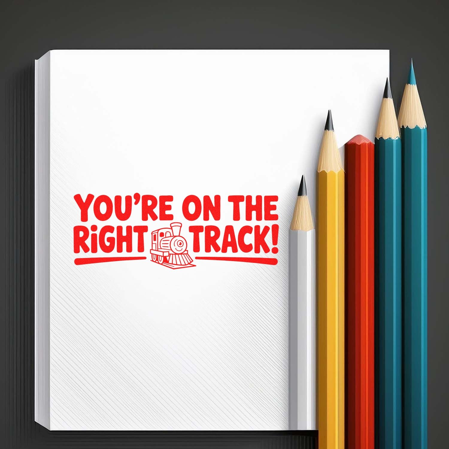 Red rubber stamp seal with train icon saying Youre on the right track! on white paper, beside colorful pencils.