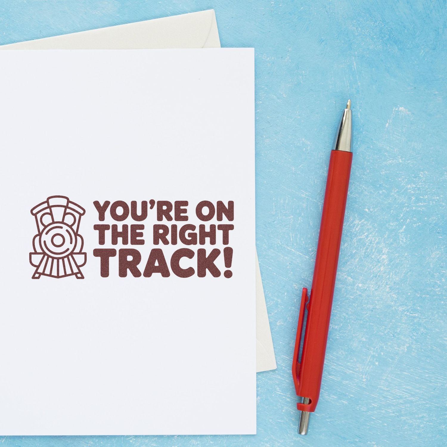 Red self-inking stamp and seal pen beside a white card with dark brown YOURE ON THE RIGHT TRACK! stamp on blue background.