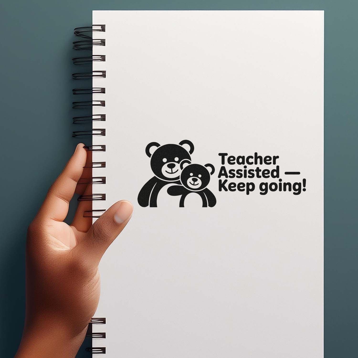 Black teacher-assist bear stamp seal on white spiral notebook, held by hand against a teal background.