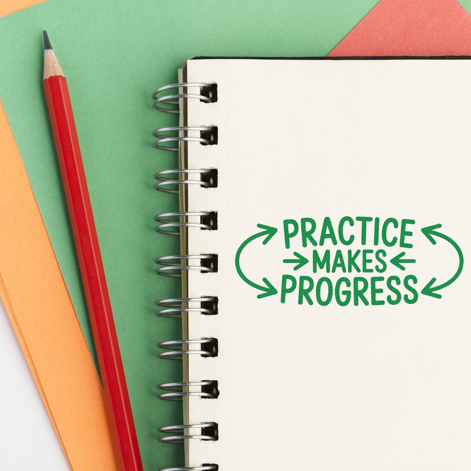 Green motivational stamp/seal reading Practice makes progress on a spiral notebook, with a red pencil and colorful paper.