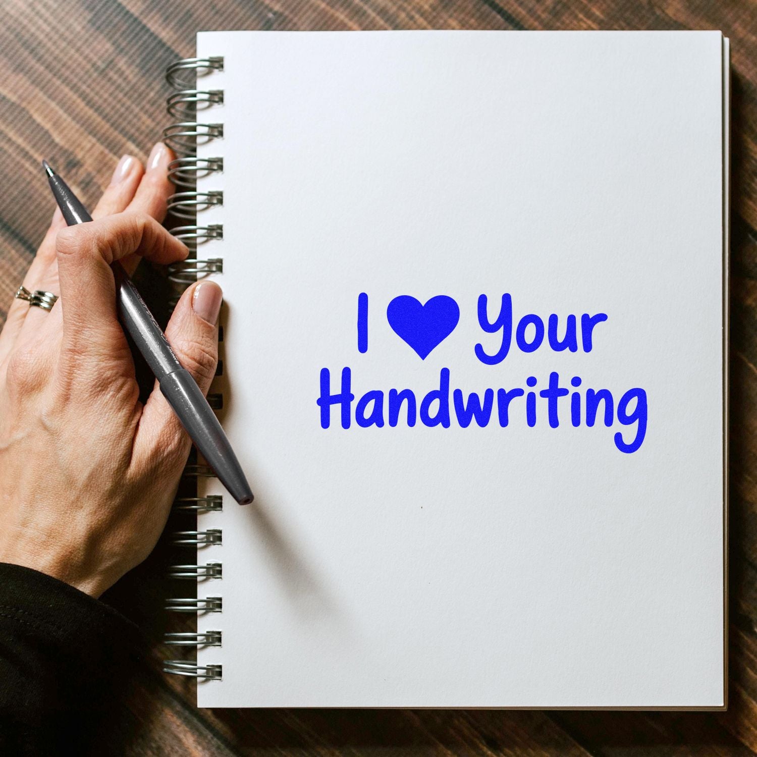 Blue custom stamp reading I ? Your Handwriting on white spiral notebook, held with a black pen.