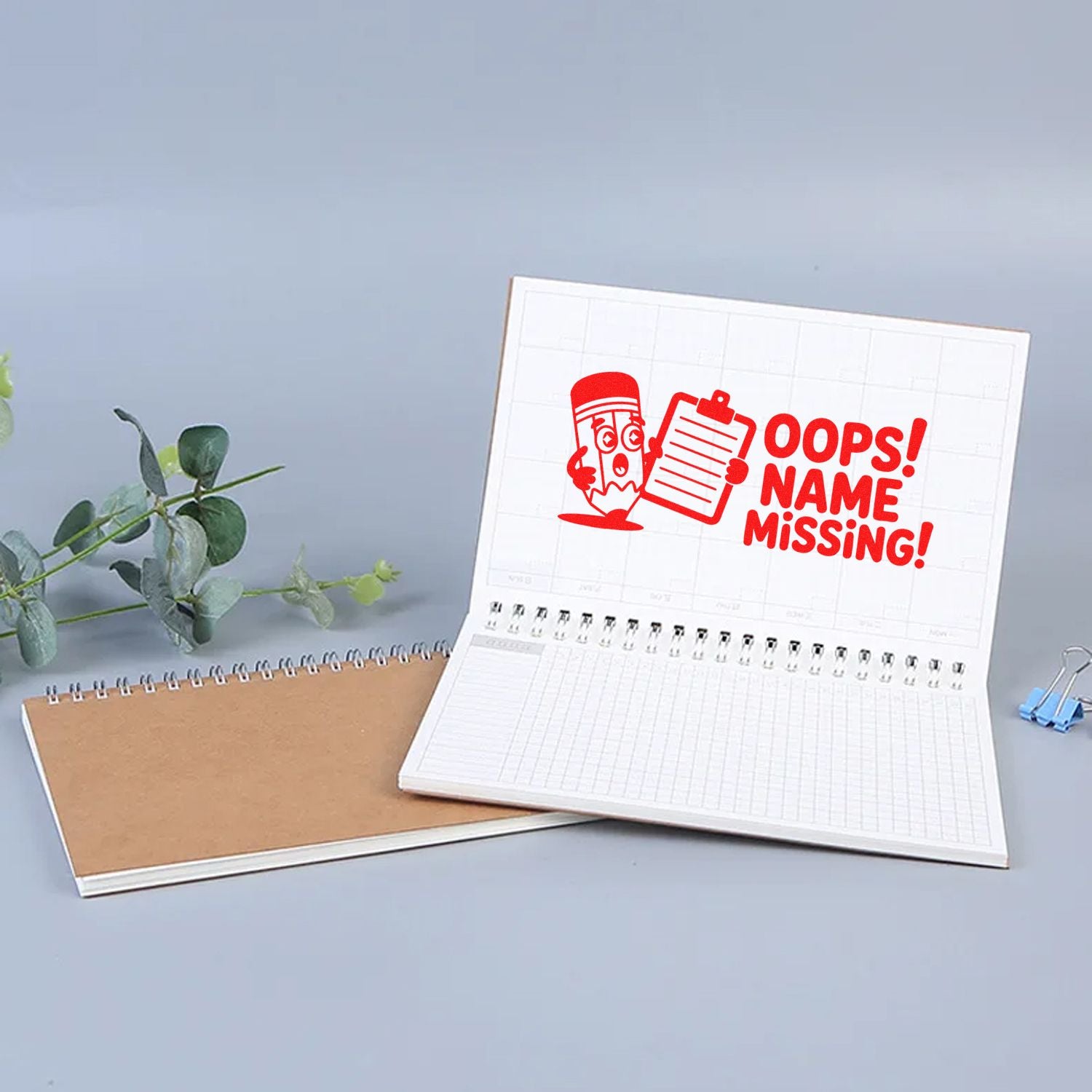 White spiral notebook with red OOPS! NAME MISSING! stamp seal, plus brown notebook and blue binder clip.