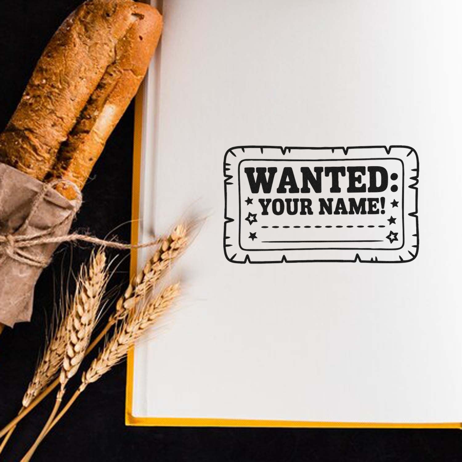 Custom black WANTED: YOUR NAME! stamp on white paper, beside a rustic bread loaf and wheat stalks.