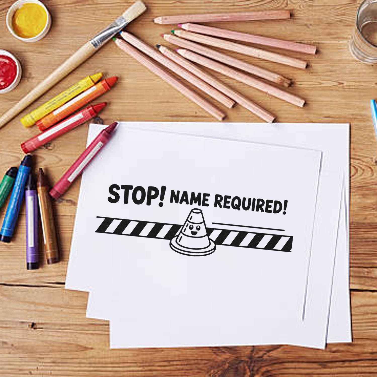 Custom stamp and seal design: black STOP! NAME REQUIRED! on white paper, with colored pencils on a wood table.