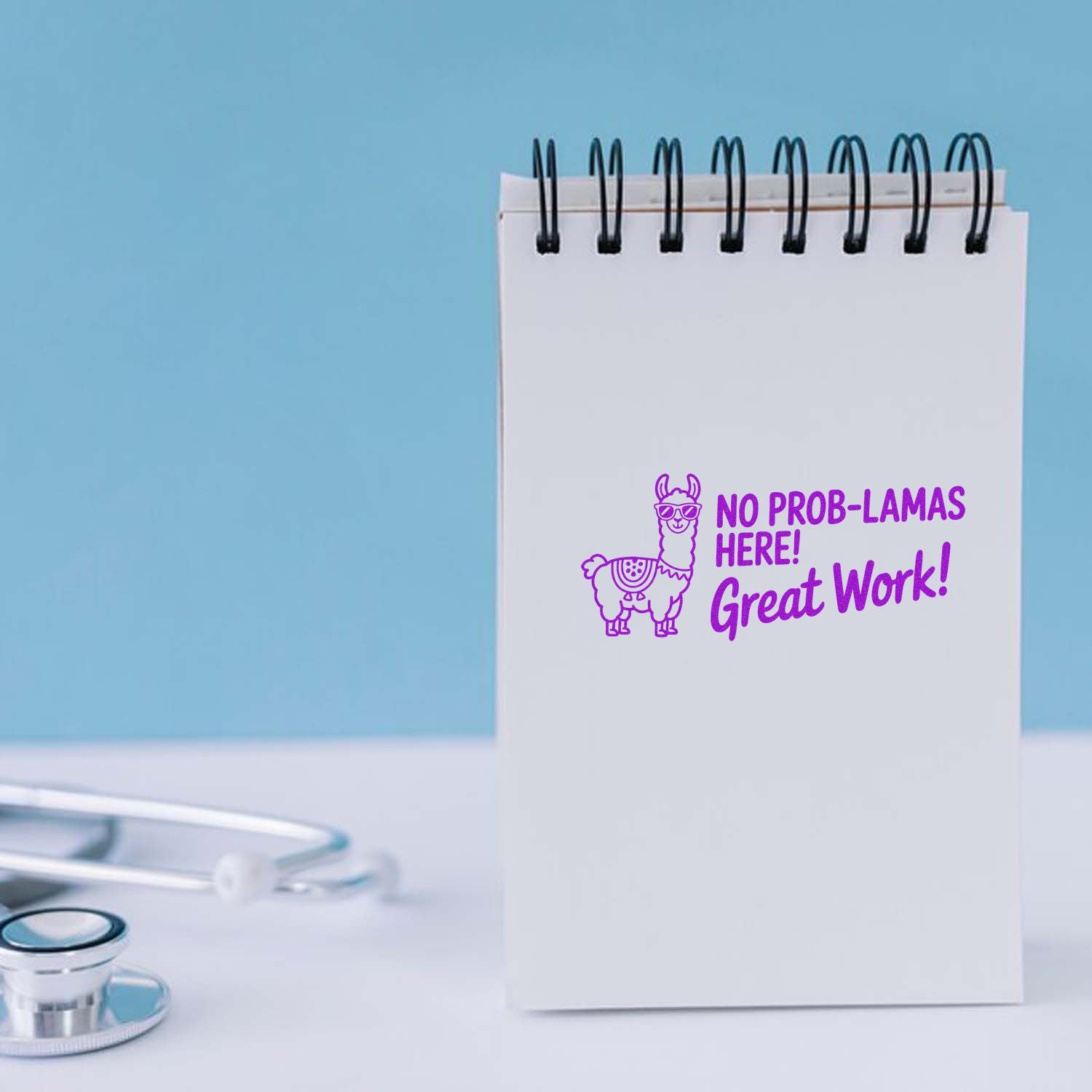 White spiral notepad with purple llama stamp/seal reading No prob-lamas here! Great Work! on a light blue desk.