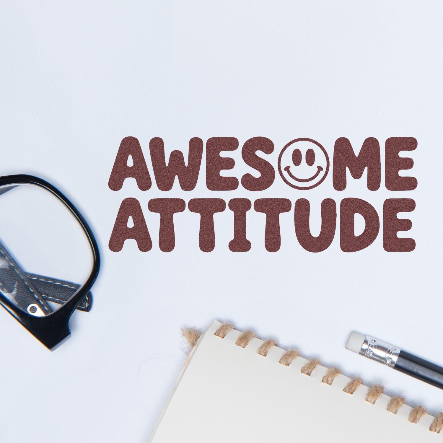 Brown stamp and seal reading AWESOME ATTITUDE with smiley face, on white paper beside notebook, pen and black clip.