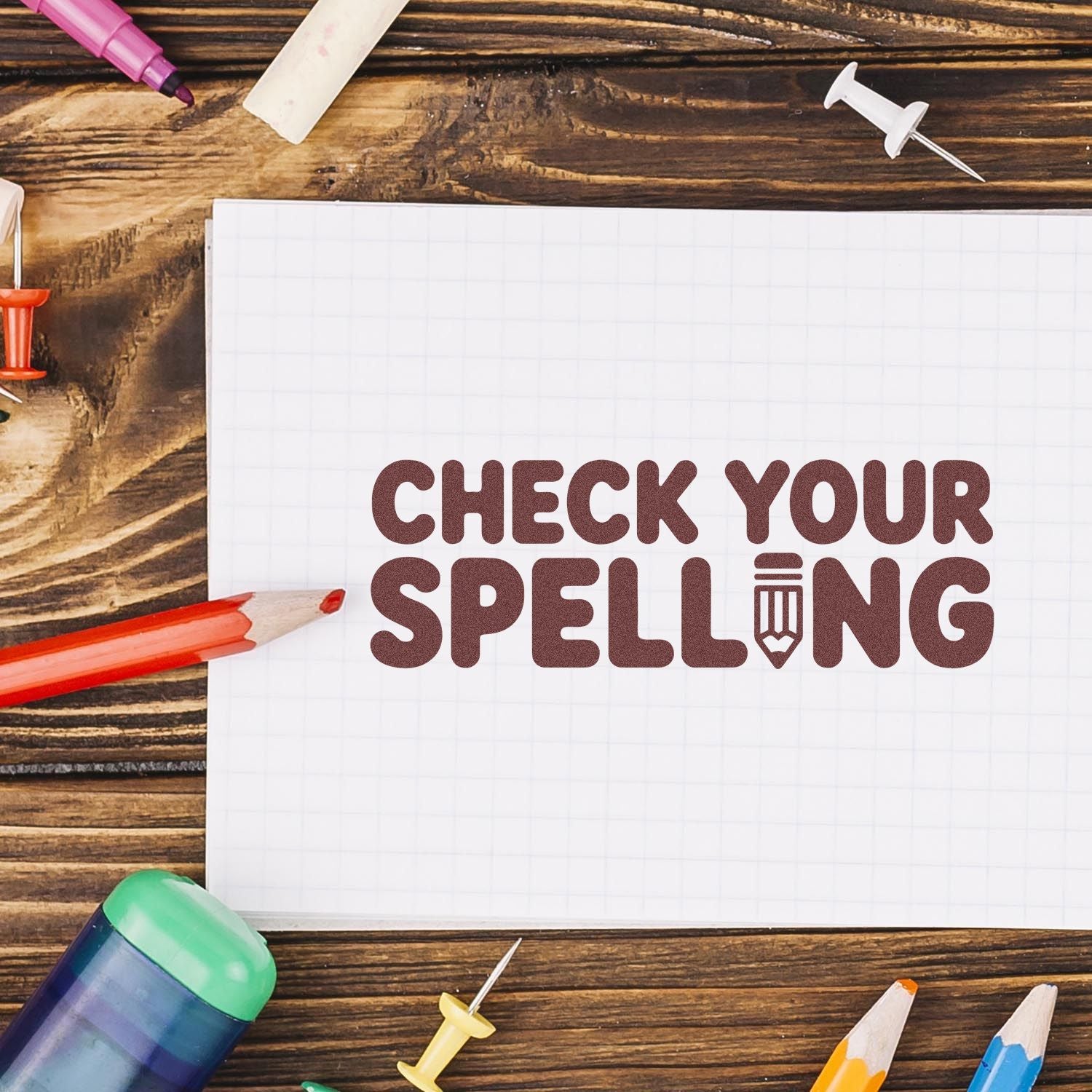 Brown Check Your Spelling stamp on white grid paper, with colorful pencils and stamp-and-seal supplies on a wood desk.