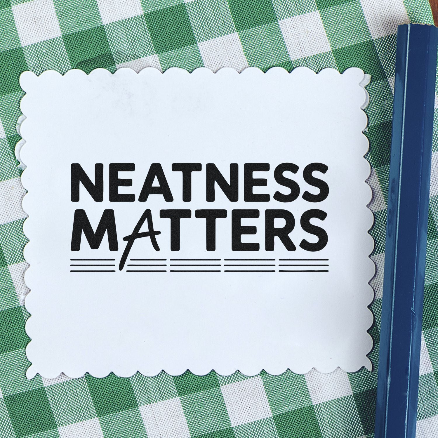 White scalloped seal stamp with black NEATNESS MATTERS text on green checkered background, beside a blue stamp tool.