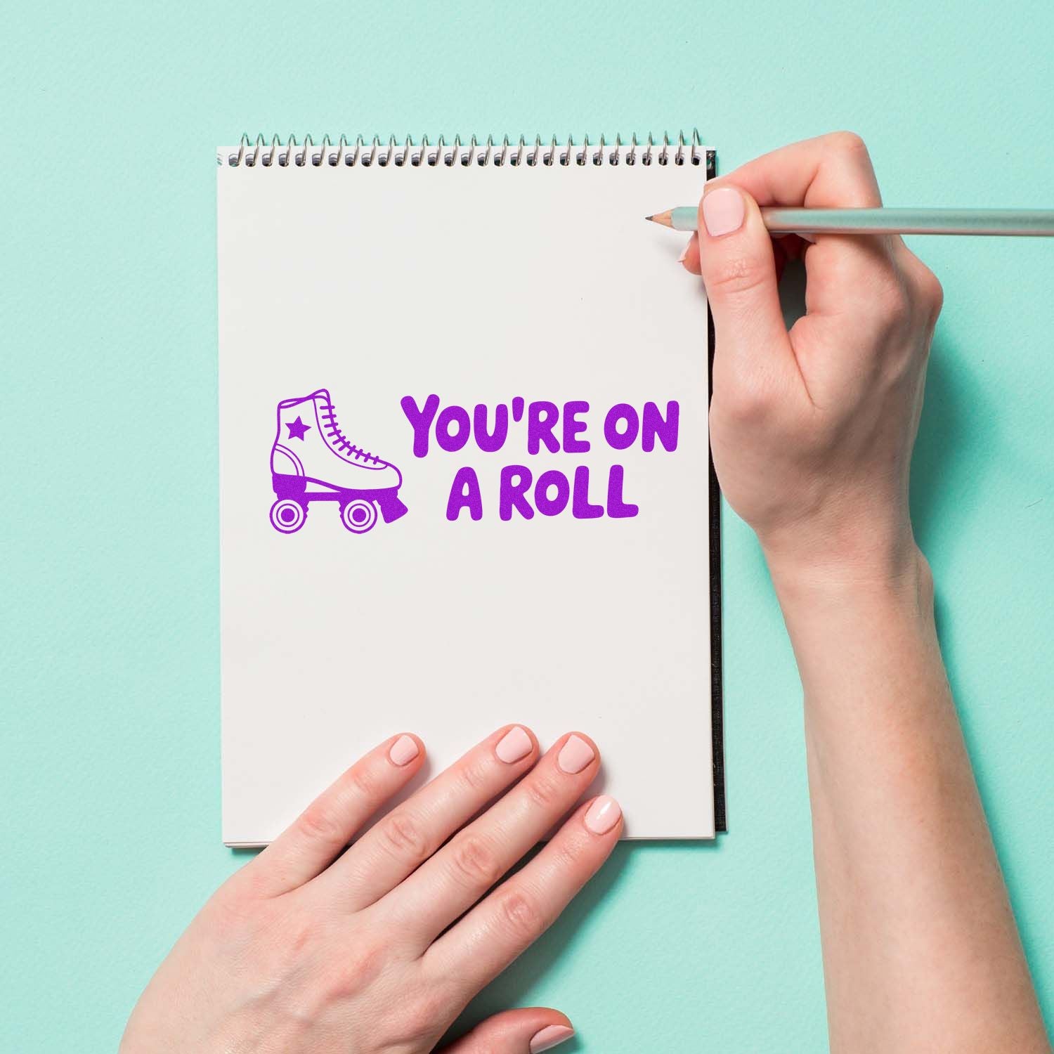 Purple roller skate stamp with Youre on a Roll on a white spiral notebook, held with a pencil on a mint background.