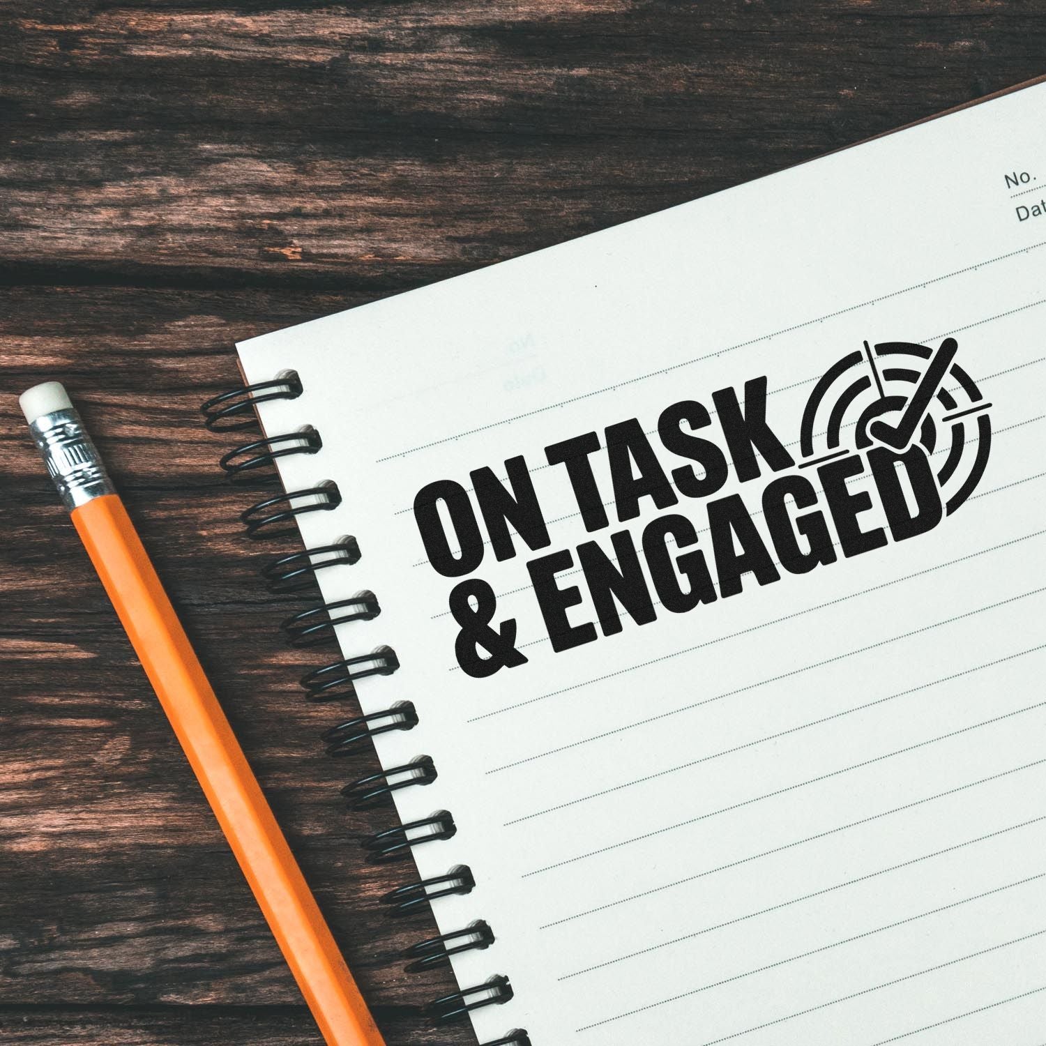 Black ON TASK & ENGAGED stamp-and-seal design on white spiral notebook, with orange pencil on dark wood desk.