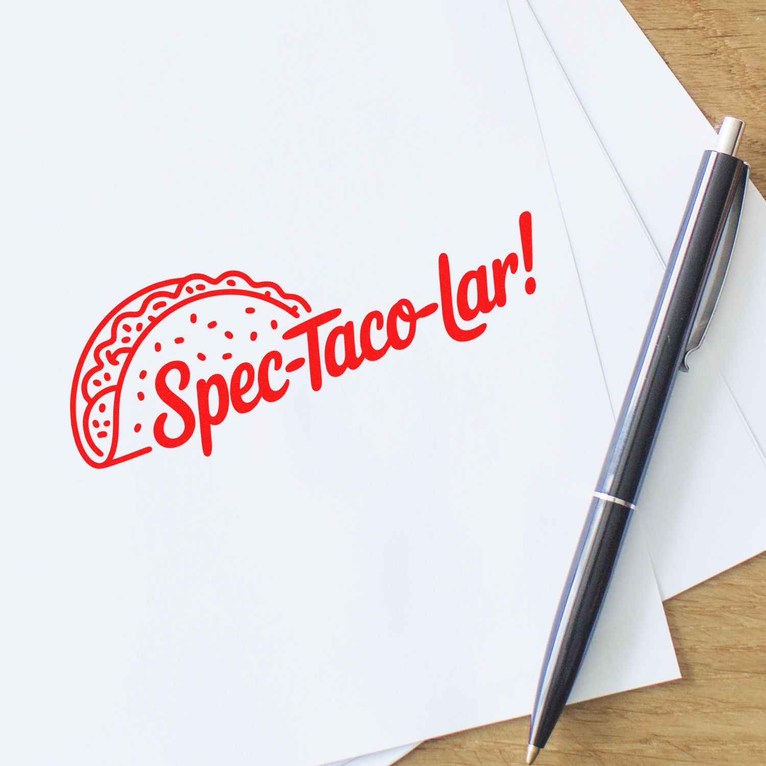 Red taco-shaped custom stamp seal reading Spec-Taco-lar! on white paper, with black pen on a wood desk.