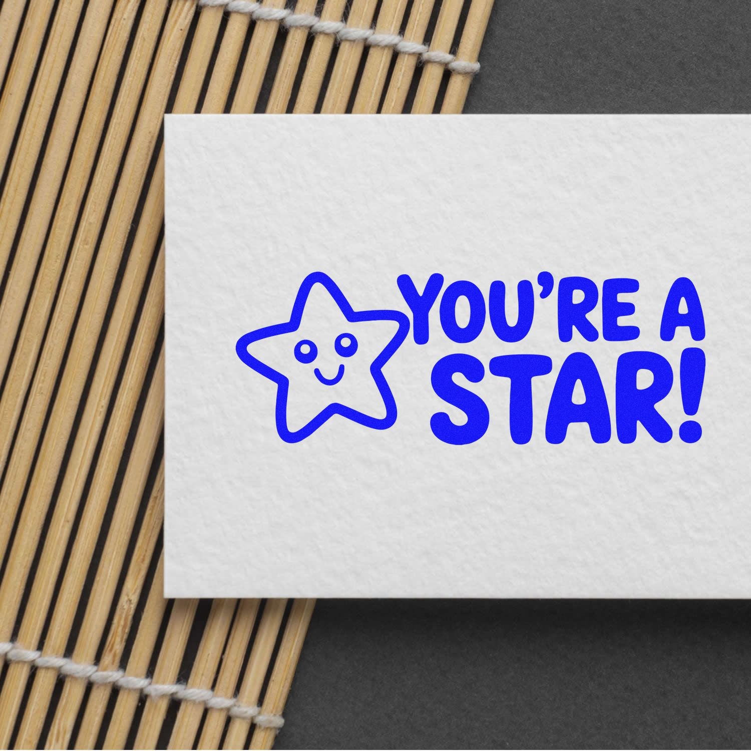 Blue Youre a Star! stamp and seal design with a smiling star on white card over a bamboo mat.
