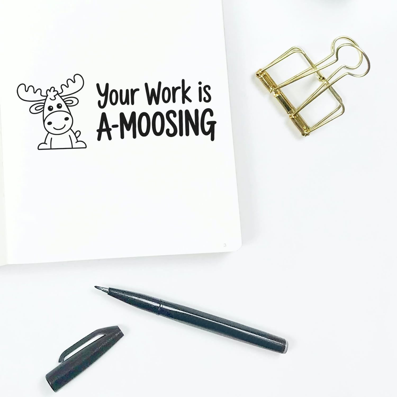 Moose-themed rubber stamp reading Your Work is A-moosing, beside a gold binder clip and black pen on white paper.