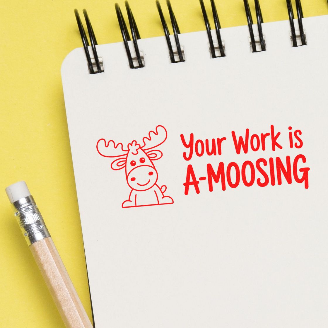 White spiral notebook on yellow background with red moose stamp seal reading Your Work is A-MOOSING beside a pencil.