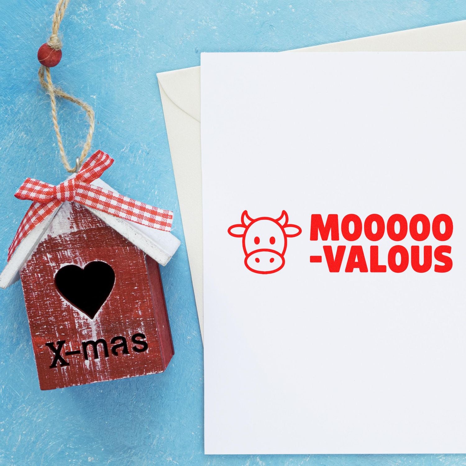 Red wooden birdhouse seal ornament with heart cutout beside white card stamped in red with cow icon and MOOO-VALOUS on blue background.