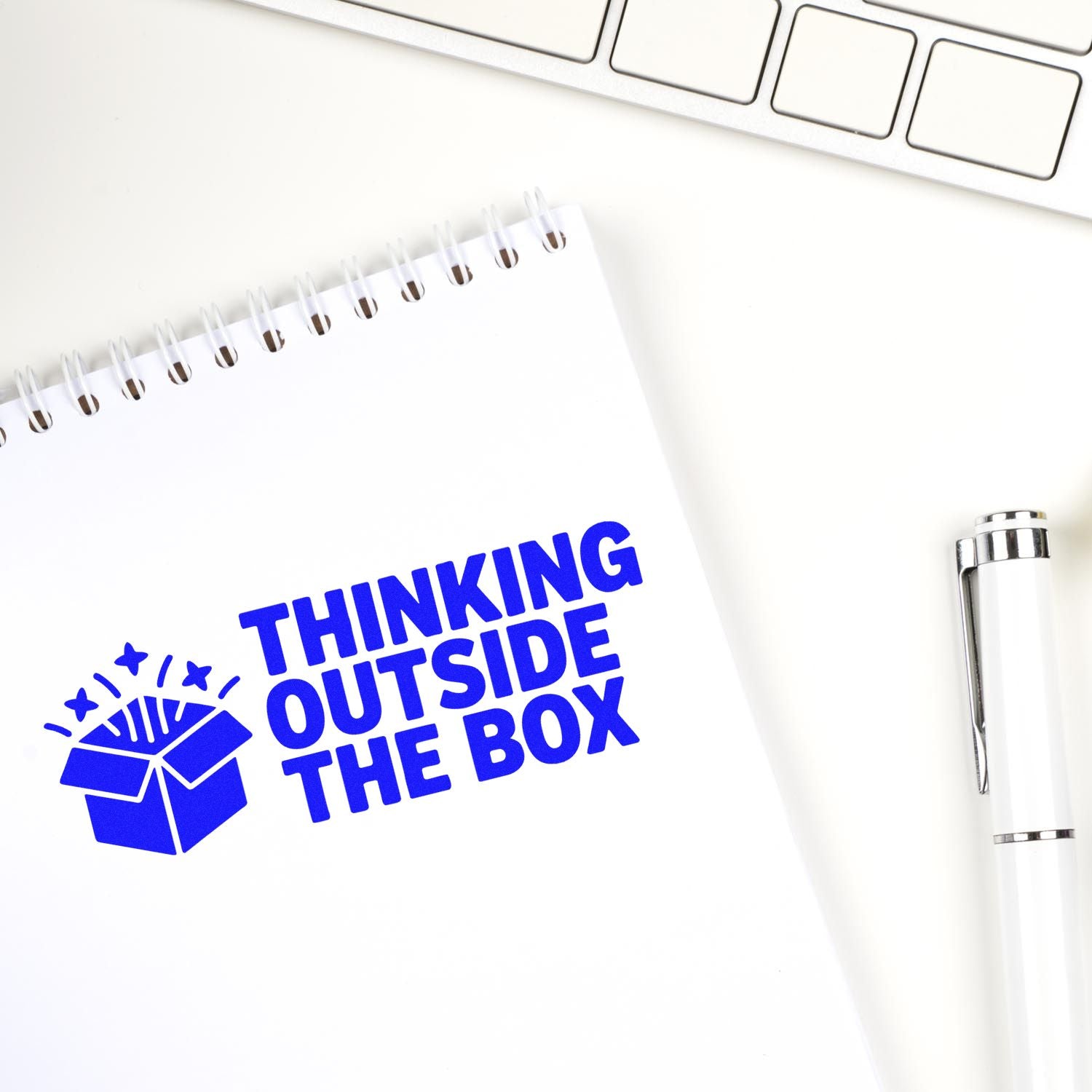 Blue custom stamp seal reading Thinking Outside the Box with open box icon on a white notepad beside a silver pen and keyboard.