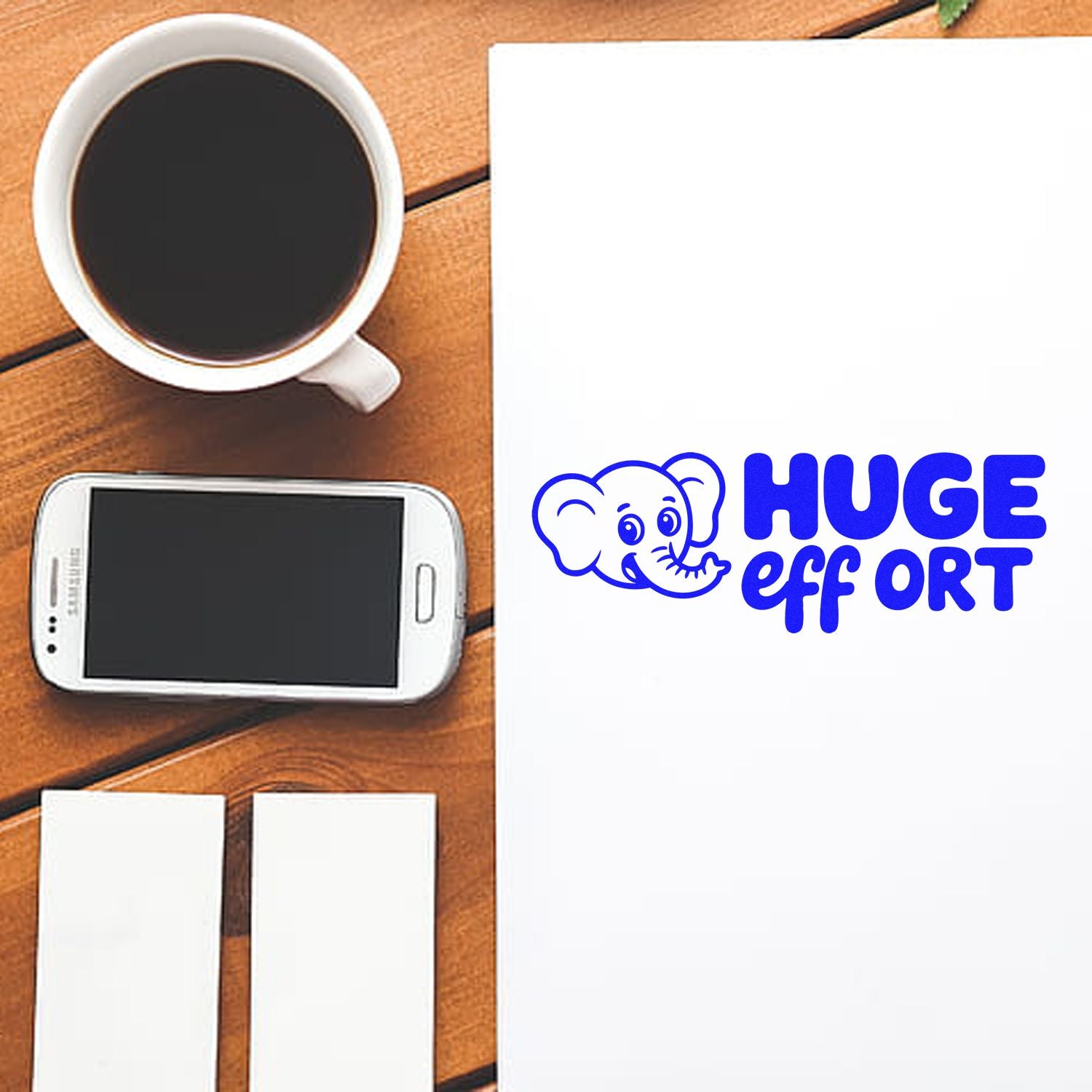 Blue elephant stamp seal with HUGE effort on white paper, beside coffee cup, phone, and blank cards on a wooden desk.