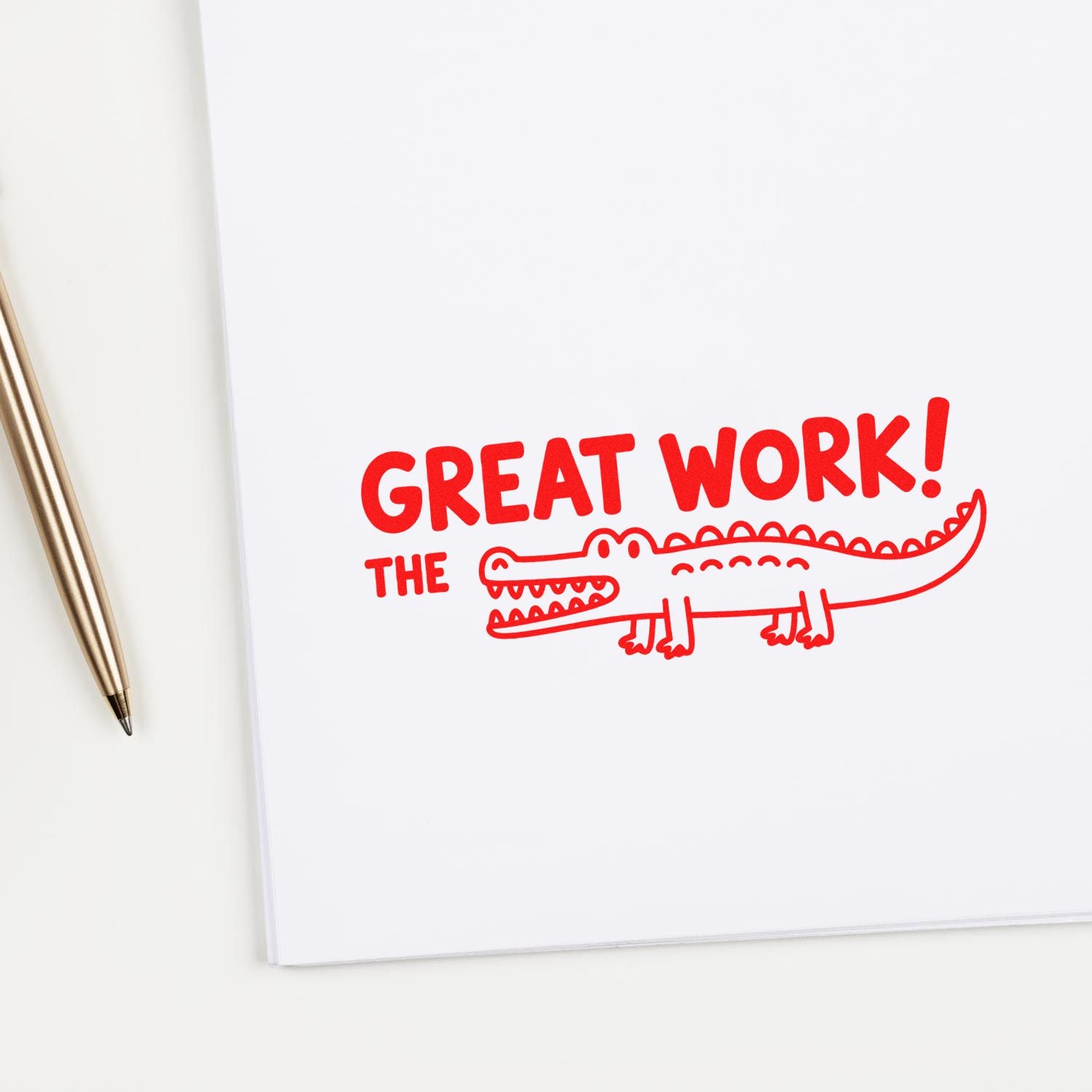 Red self-inking stamp reading GREAT WORK! THE with crocodile seal on white paper, beside a gold pen.