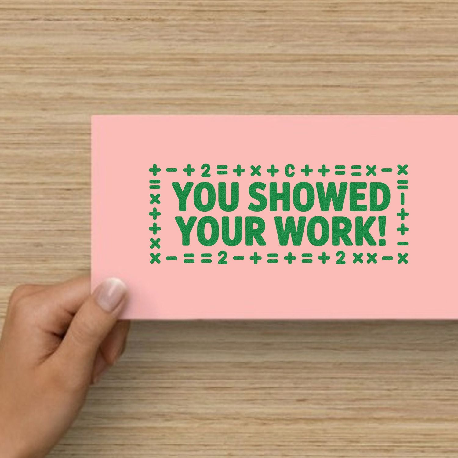 Pink stamp-and-seal card with green YOU SHOWED YOUR WORK! text and math symbols, held by hand.