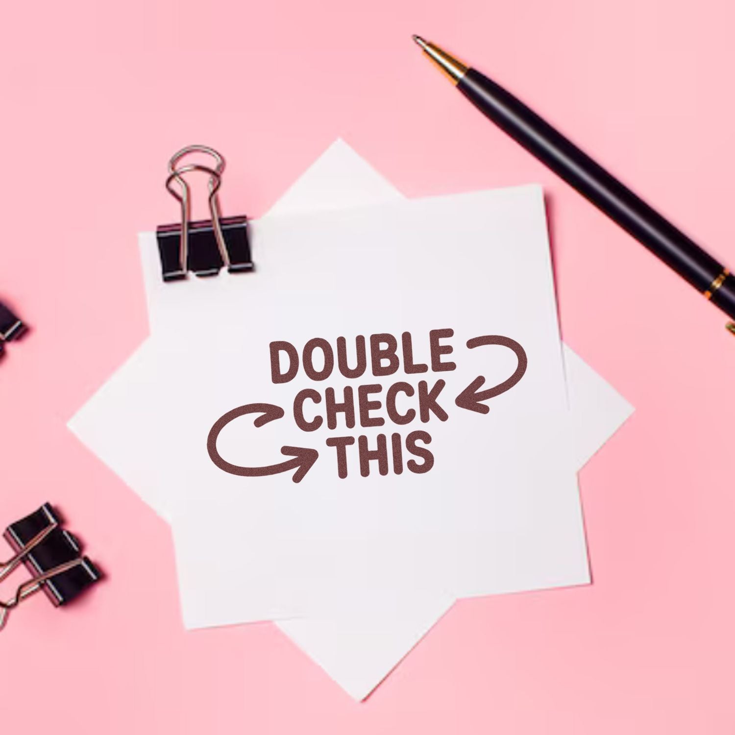 Brown DOUBLE CHECK THIS custom stamp on white paper, with black pen and binder clips on a pink background.