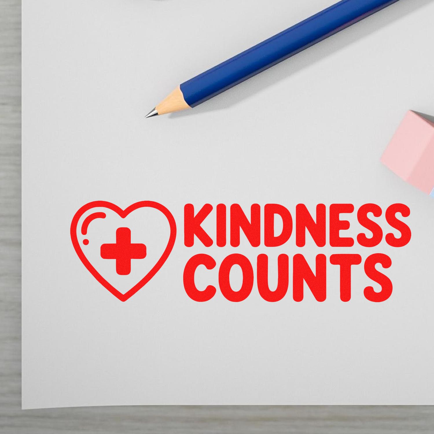 Red KINDNESS COUNTS stamp seal with heart and cross, beside a blue pencil on white paper.