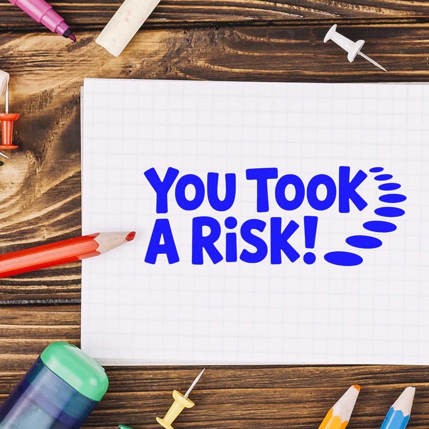 Blue You Took A Risk! on graph paper notebook, with stamp and seal tools, pencils, and pushpins on a wood table.