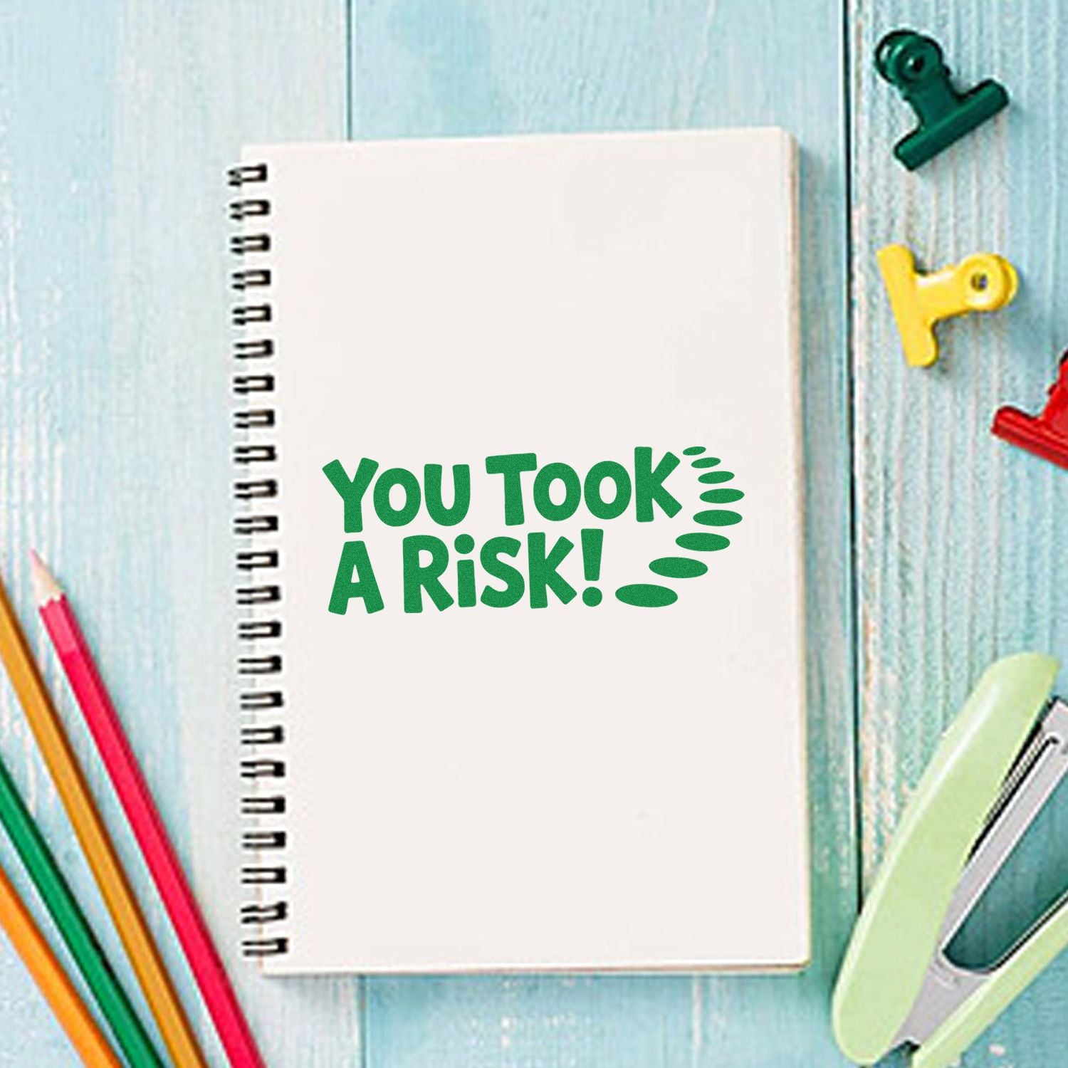 White spiral notebook with green You Took A Risk! stamp, surrounded by colorful pencils and desk stamp and seal tools.