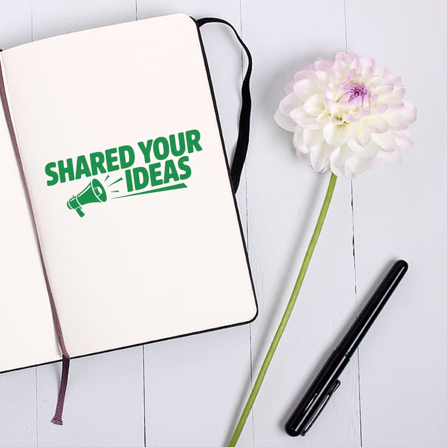 Cream notebook with green stamp seal reading Shared Your Ideas, beside a black pen and white flower.