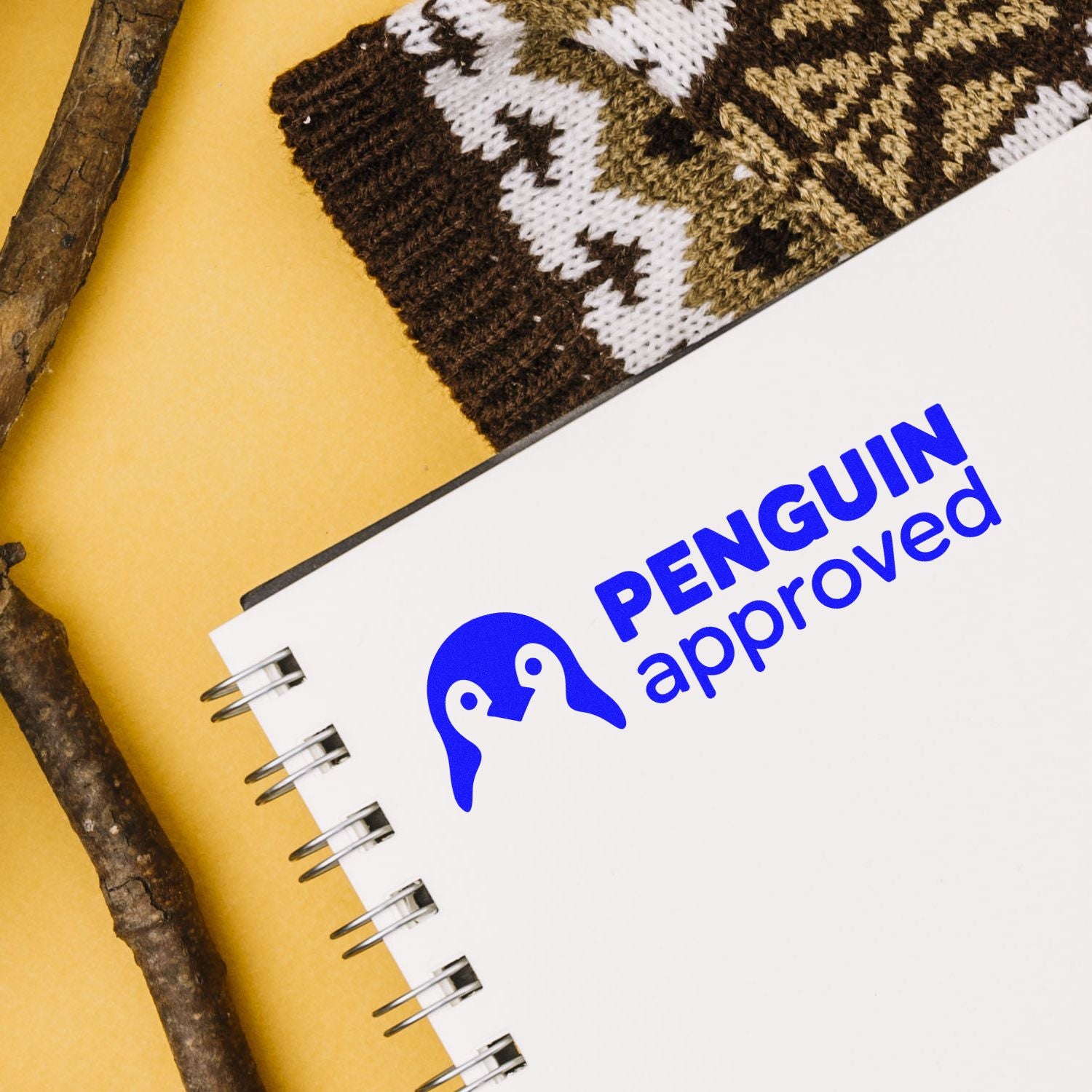 Blue Penguin Approved stamp seal on a white spiral notebook, with brown knit fabric and twigs on a yellow background.
