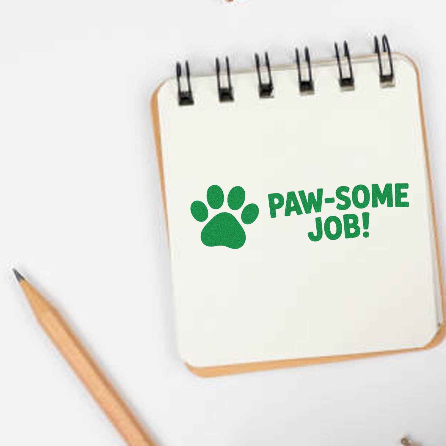 Green paw-print stamp with PAW-SOME JOB! on cream spiral notepad, with a pencil on a white background.