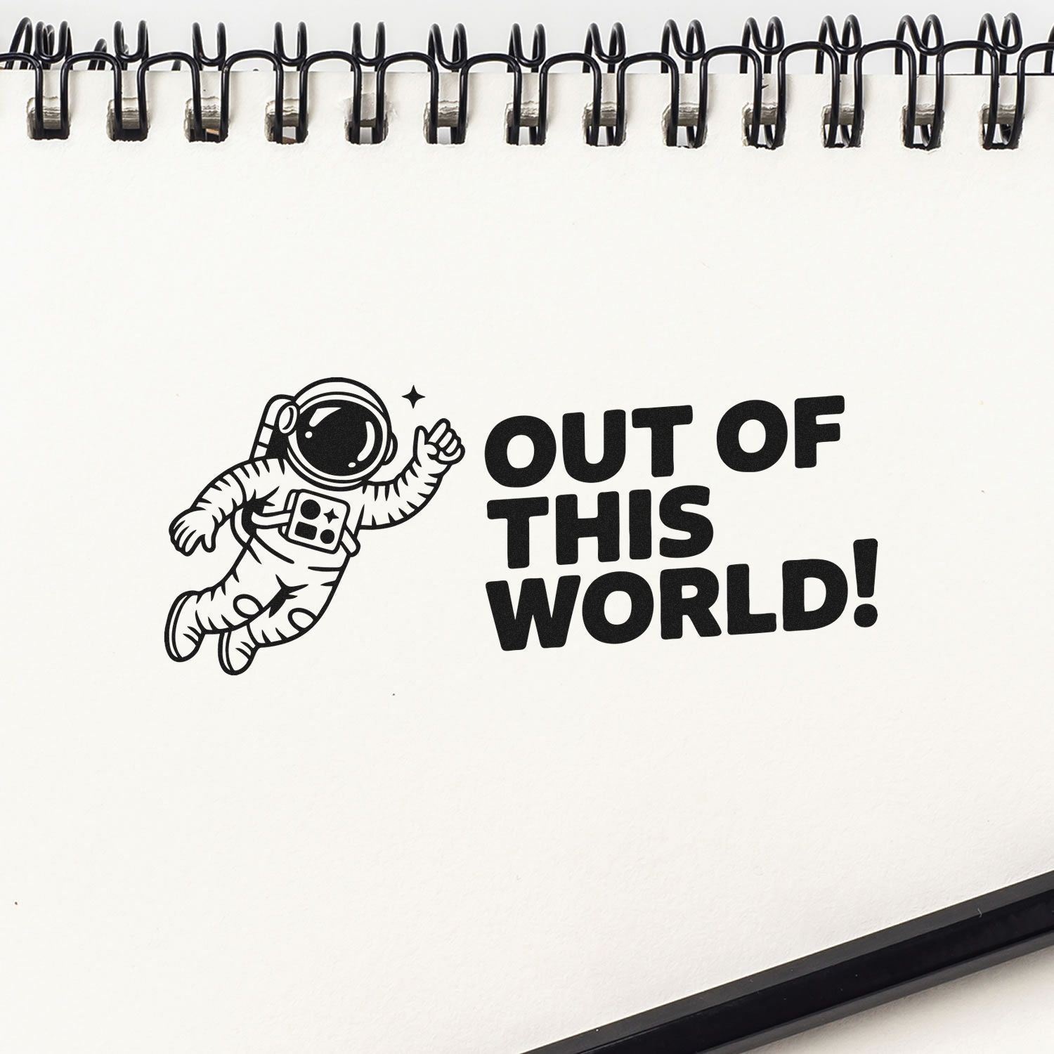 Black stamp and seal design of an astronaut with OUT OF THIS WORLD! on a white spiral-bound notebook page.