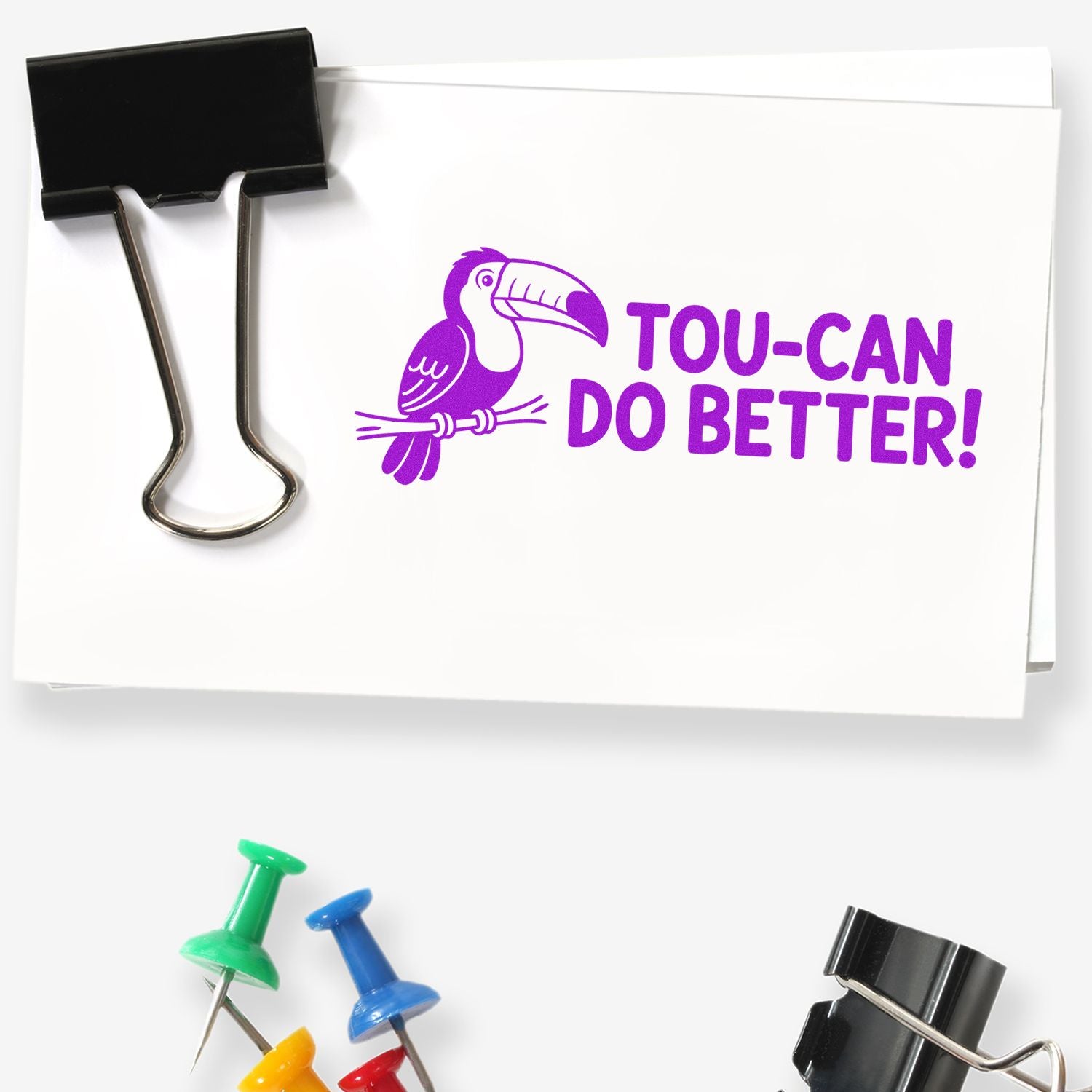 Black binder clip holding white paper stamped in purple with toucan logo and TOU-CAN DO BETTER!; colorful push pins below.