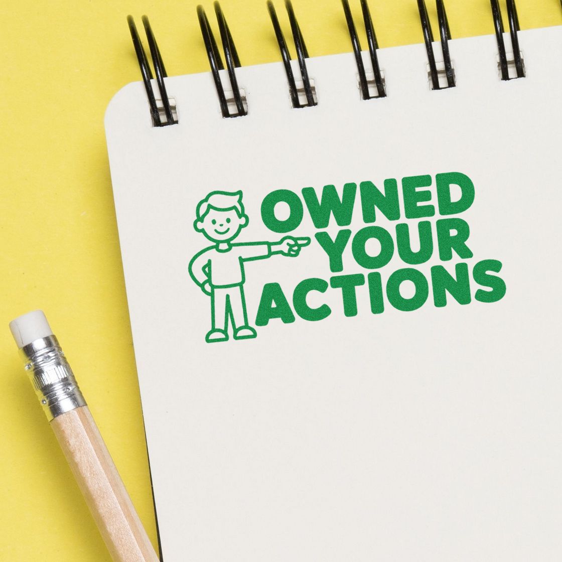 Green Owned Your Actions seal stamp with cartoon figure on white notebook, on yellow background beside a pencil.