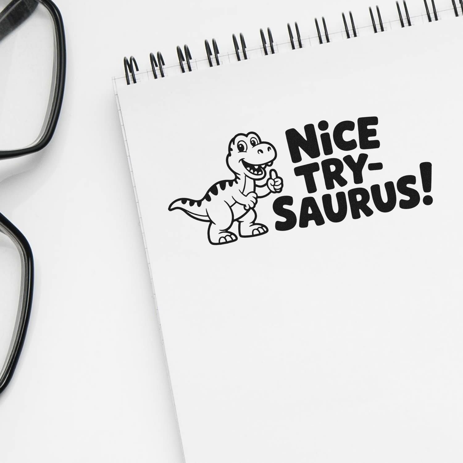Black Nice Try-saurus! stamp/seal on white spiral notepad with cartoon dinosaur; glasses edge visible.