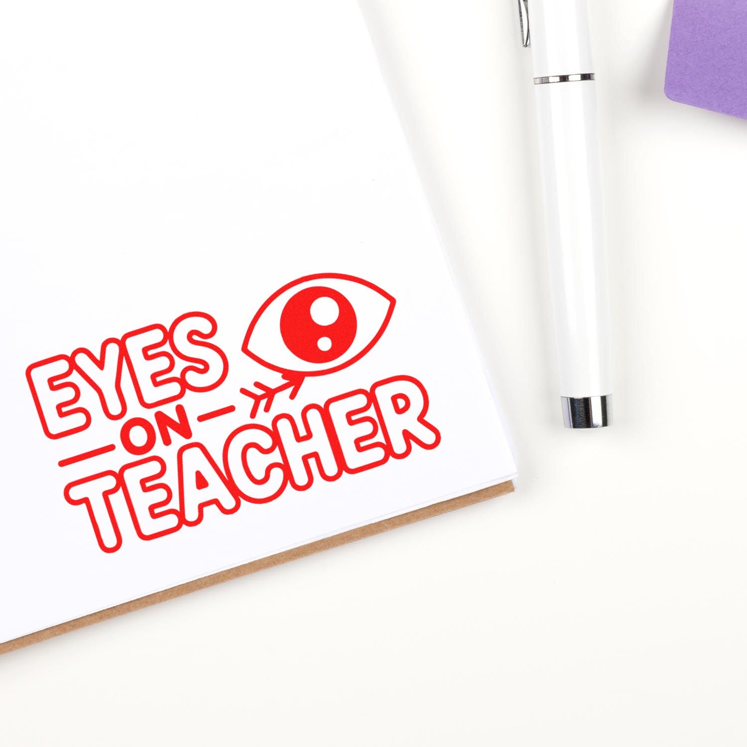 Red EYES ON TEACHER stamp seal with eye icon on white paper, beside a white pen and purple notebook.