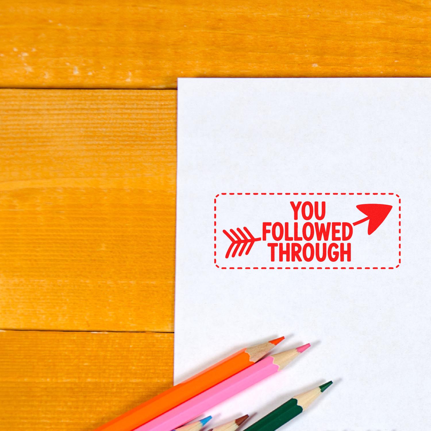 Red self-inking stamp reading You Followed Through on white paper, with colored pencils on an orange wood desk.