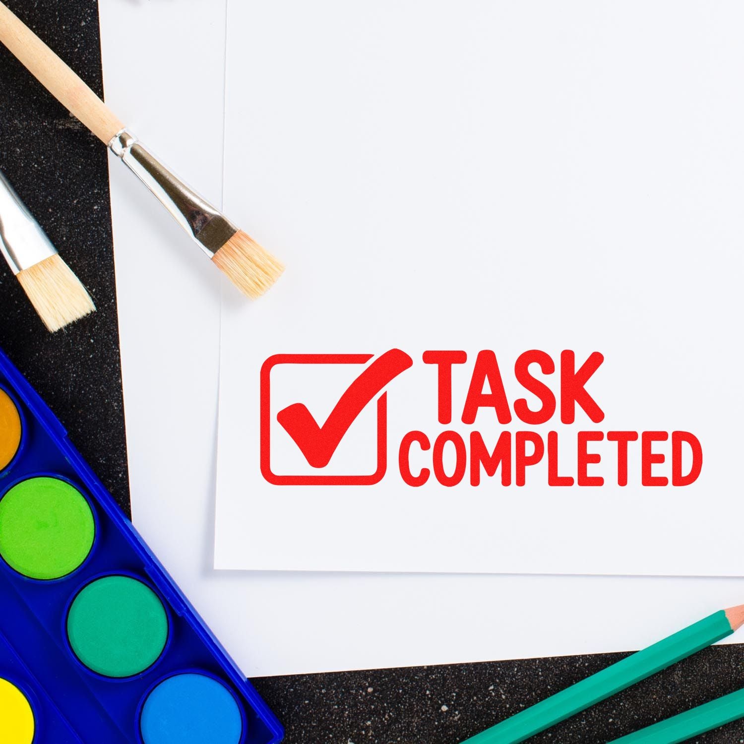 Red task completed stamp seal on white paper, with paintbrushes and colorful watercolor palette.