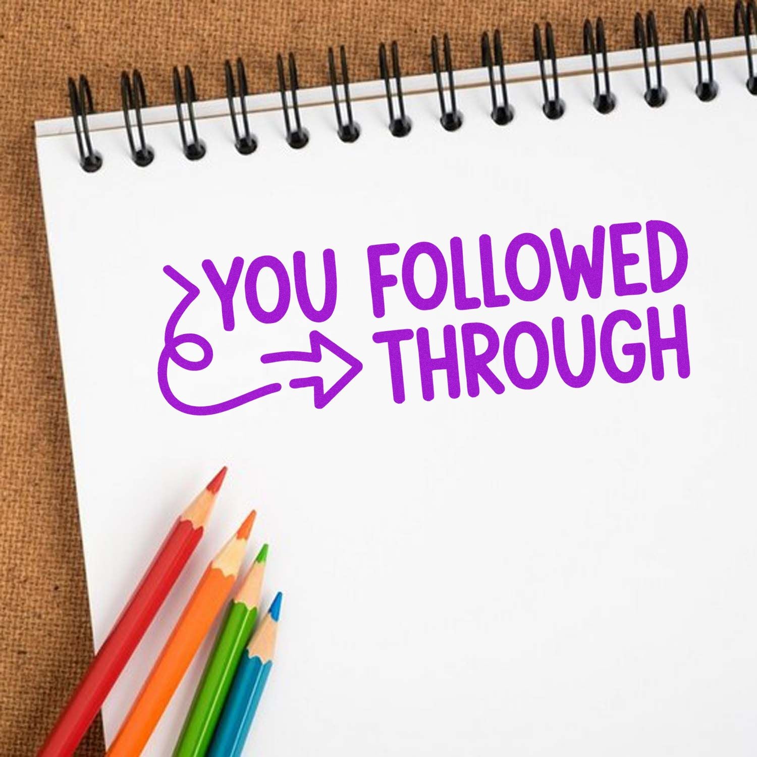 Purple YOU FOLLOWED THROUGH stamp/seal on a white spiral notebook, with colored pencils on a brown background.