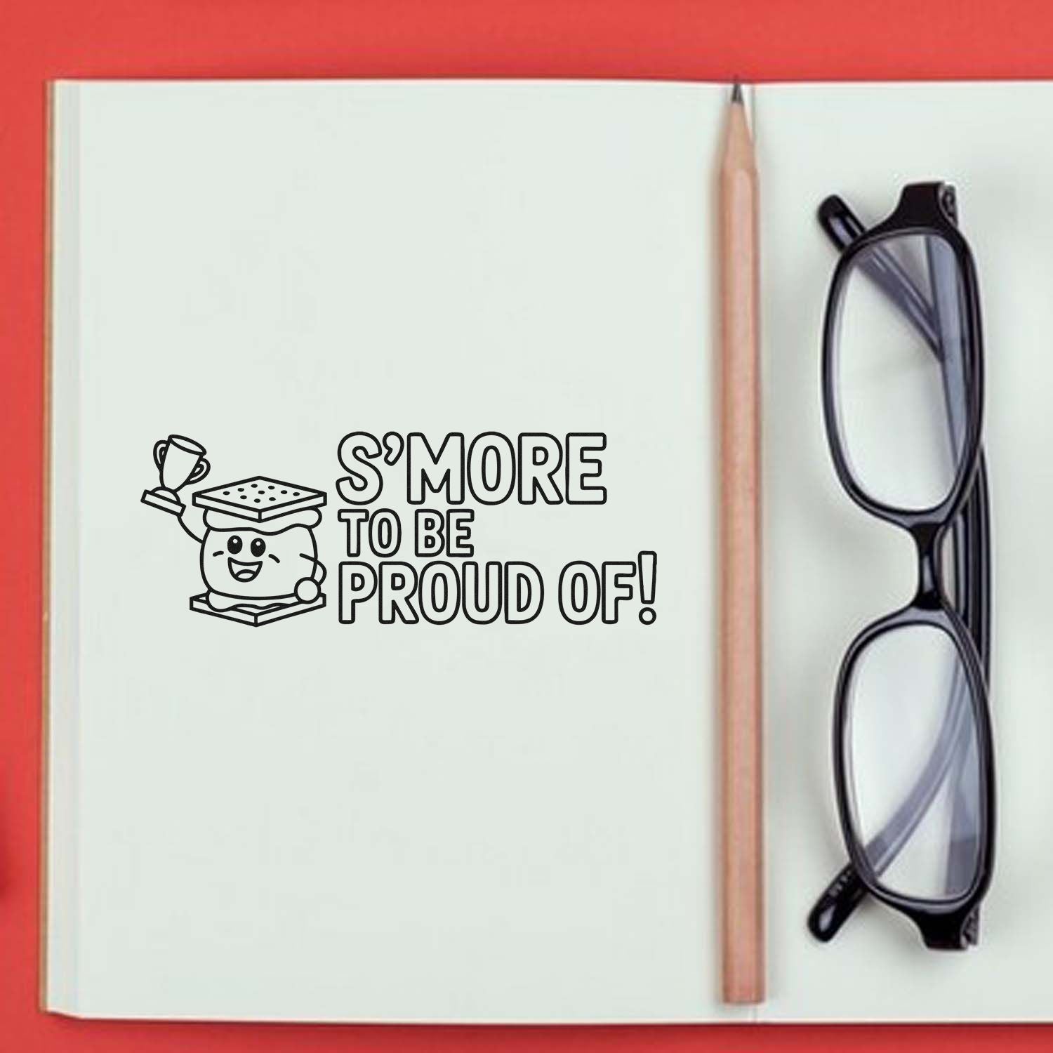 Black custom stamp and seal on an open notebook reading Smore to be proud of! with a pencil and black glasses.