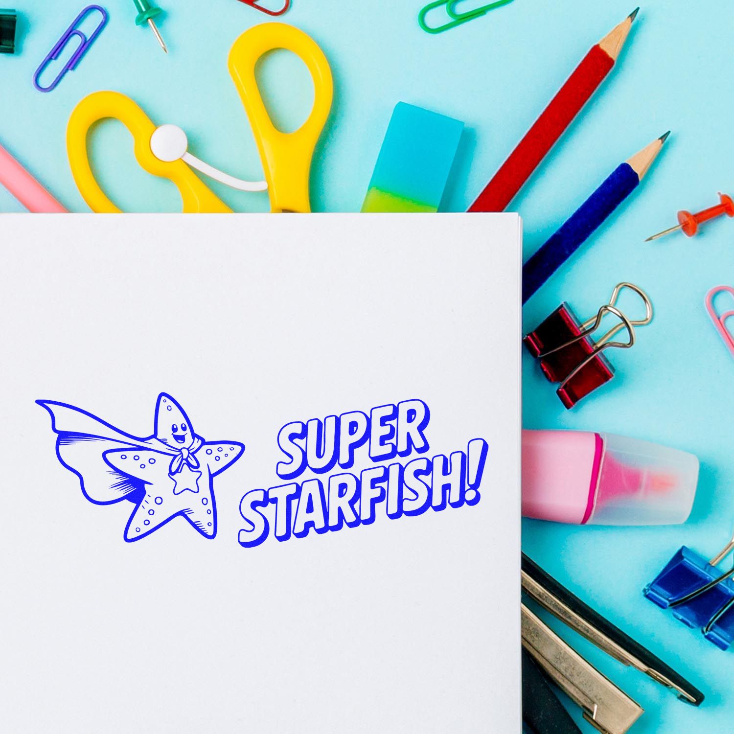 Blue SUPER STARFISH! stamp on white paper, surrounded by colorful stamp and seal products, pencils, clips, and eraser.