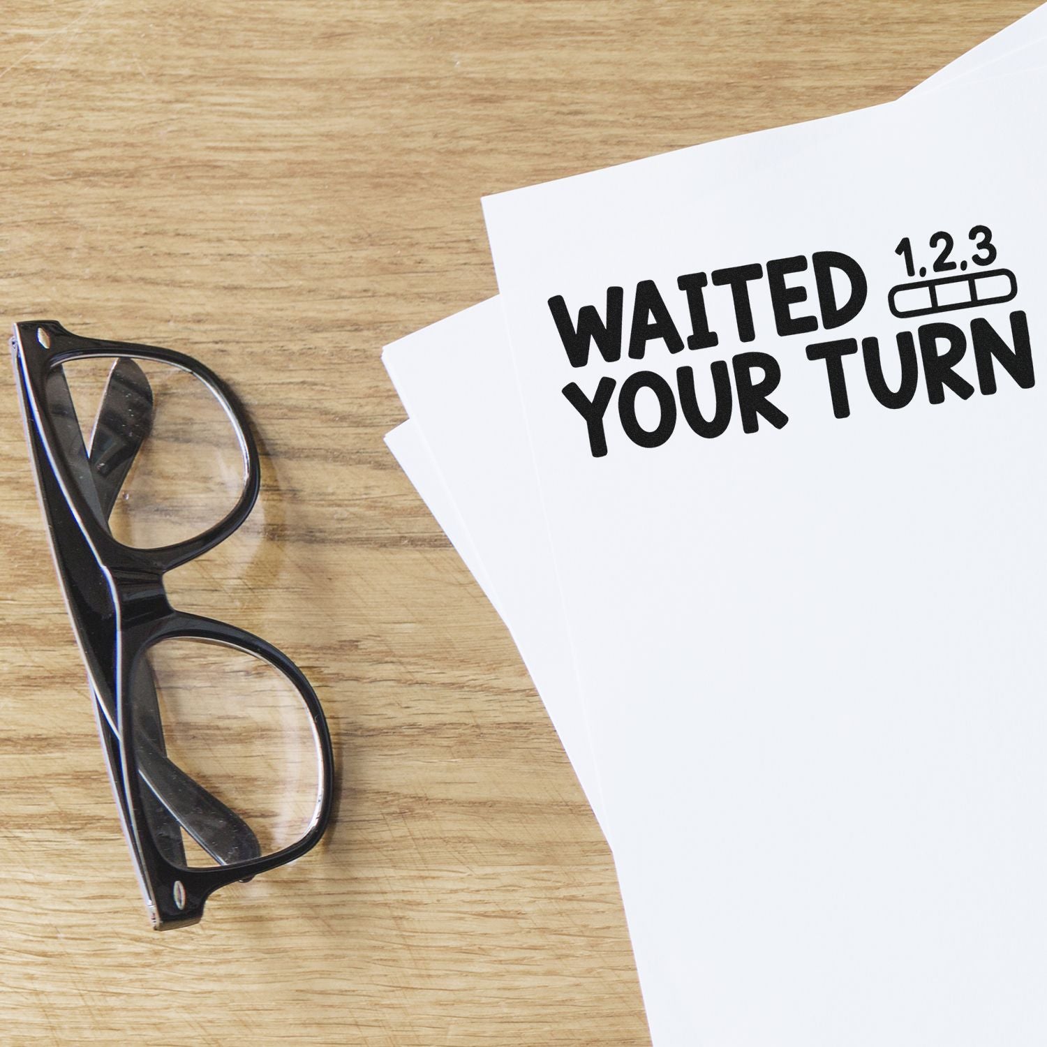 Black eyeglasses beside white stamp and seal product sheet reading WAITED YOUR TURN with numbered queue icon on wood desk.
