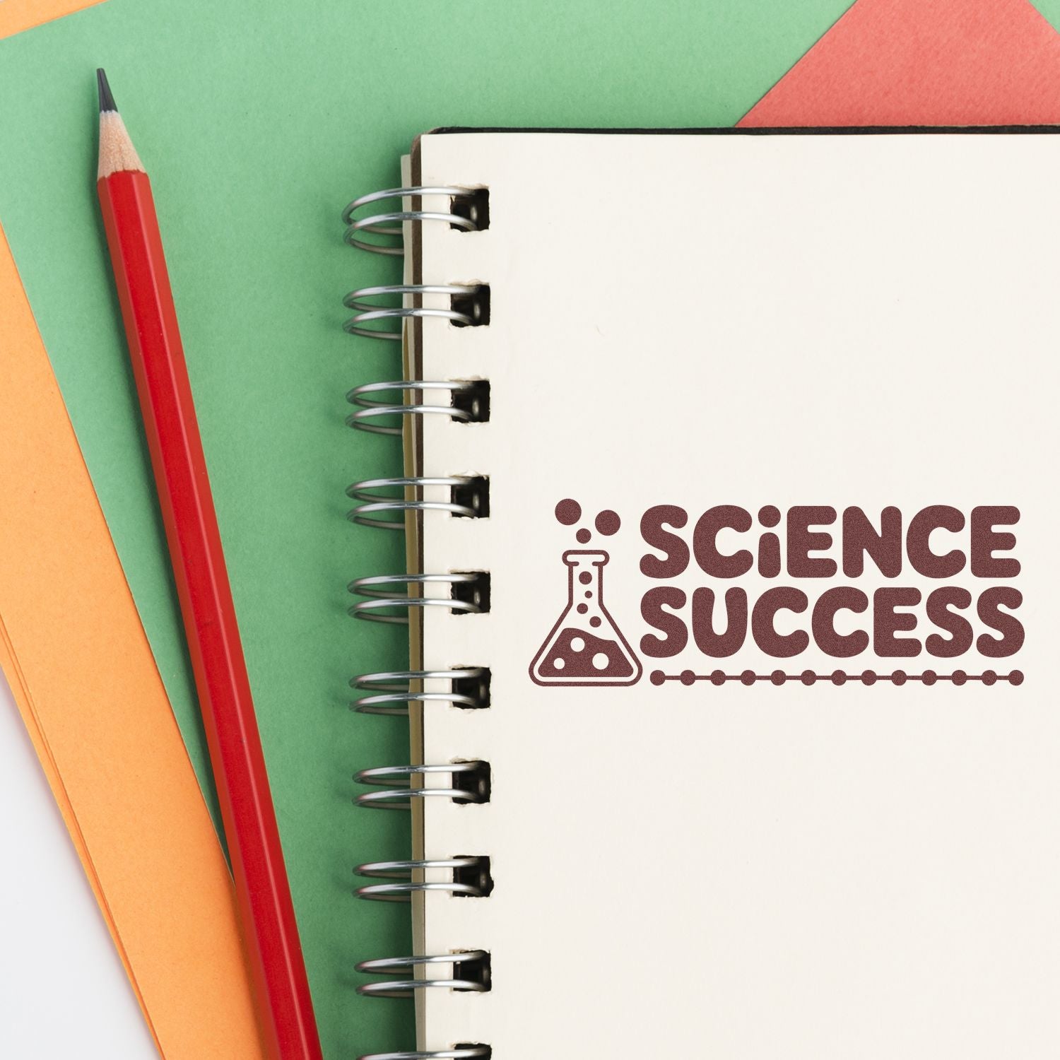 Brown Science Success stamp seal on white spiral notebook, with red pencil on green, orange, and coral paper.