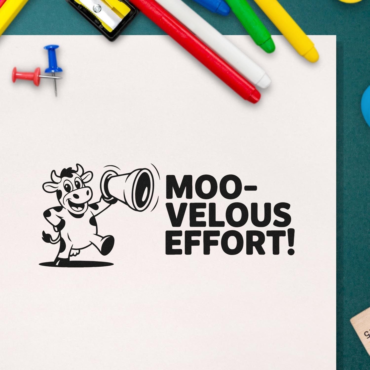 Black custom rubber stamp seal with cow mascot and MOO-VELOUS EFFORT! text, beside colorful pens and pushpins.