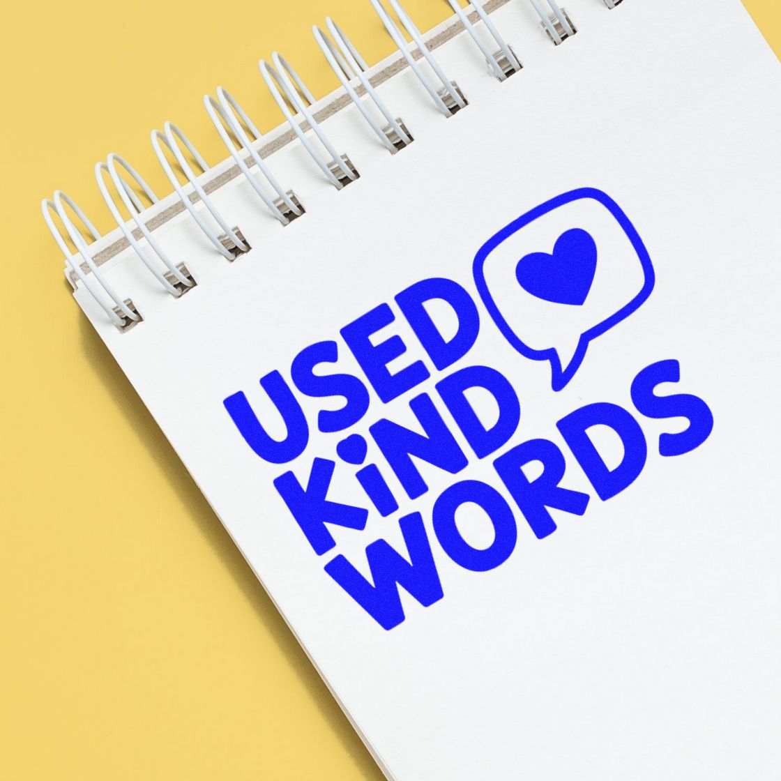 Blue USED KIND WORDS stamp and seal with heart speech bubble on a white spiral notebook, set on a yellow background.