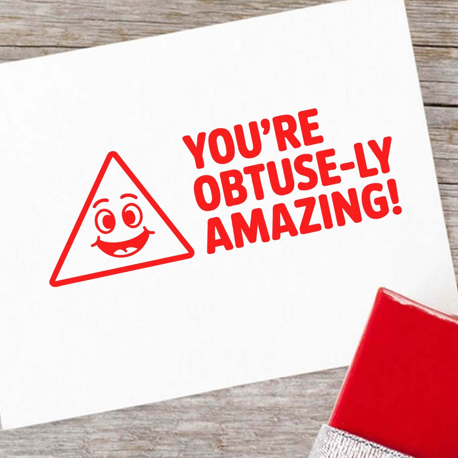 Red rubber stamp seal on white card reading YOURE OBTUSE-LY AMAZING! with a smiling triangle face.